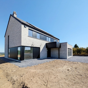 Facade avant d'une maison moderne en briques blanches avec toiture noire, sous un ciel bleu, terrain non aménagé.